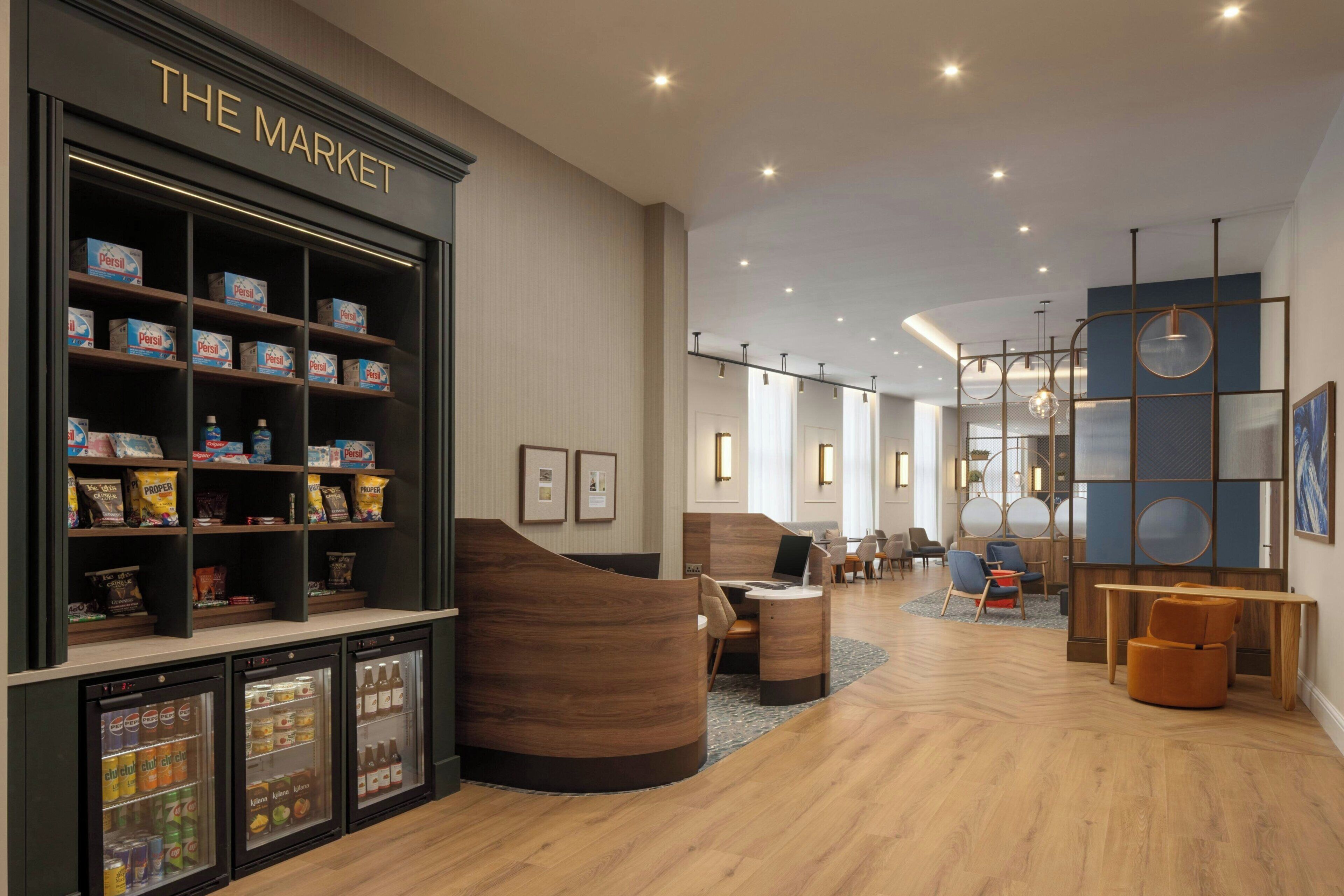 Um saguão de hotel moderno com uma área de lanches 'The Market', assentos e uma mesa de recepção.