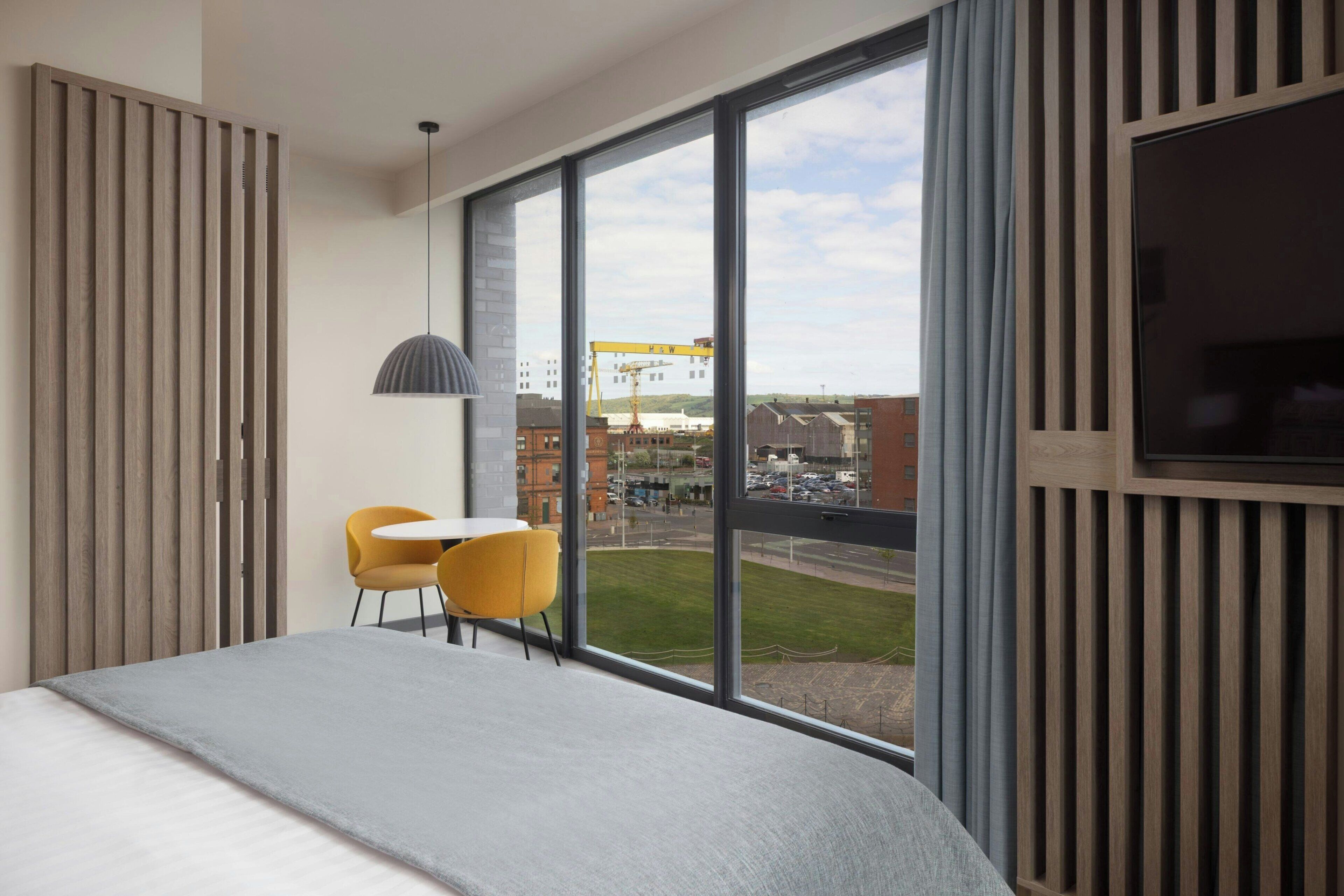 Une chambre d’hôtel avec un grand lit, une petite table avec une chaise, une télévision et une vue sur le paysage urbain grâce à de grandes fenêtres.