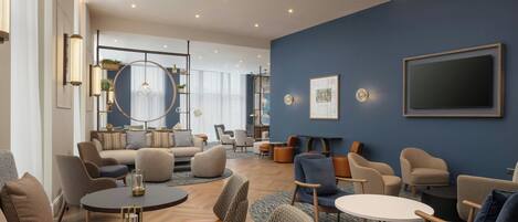 A modern hotel lobby with a seating area, a TV mounted on the wall, and a large window providing natural light.