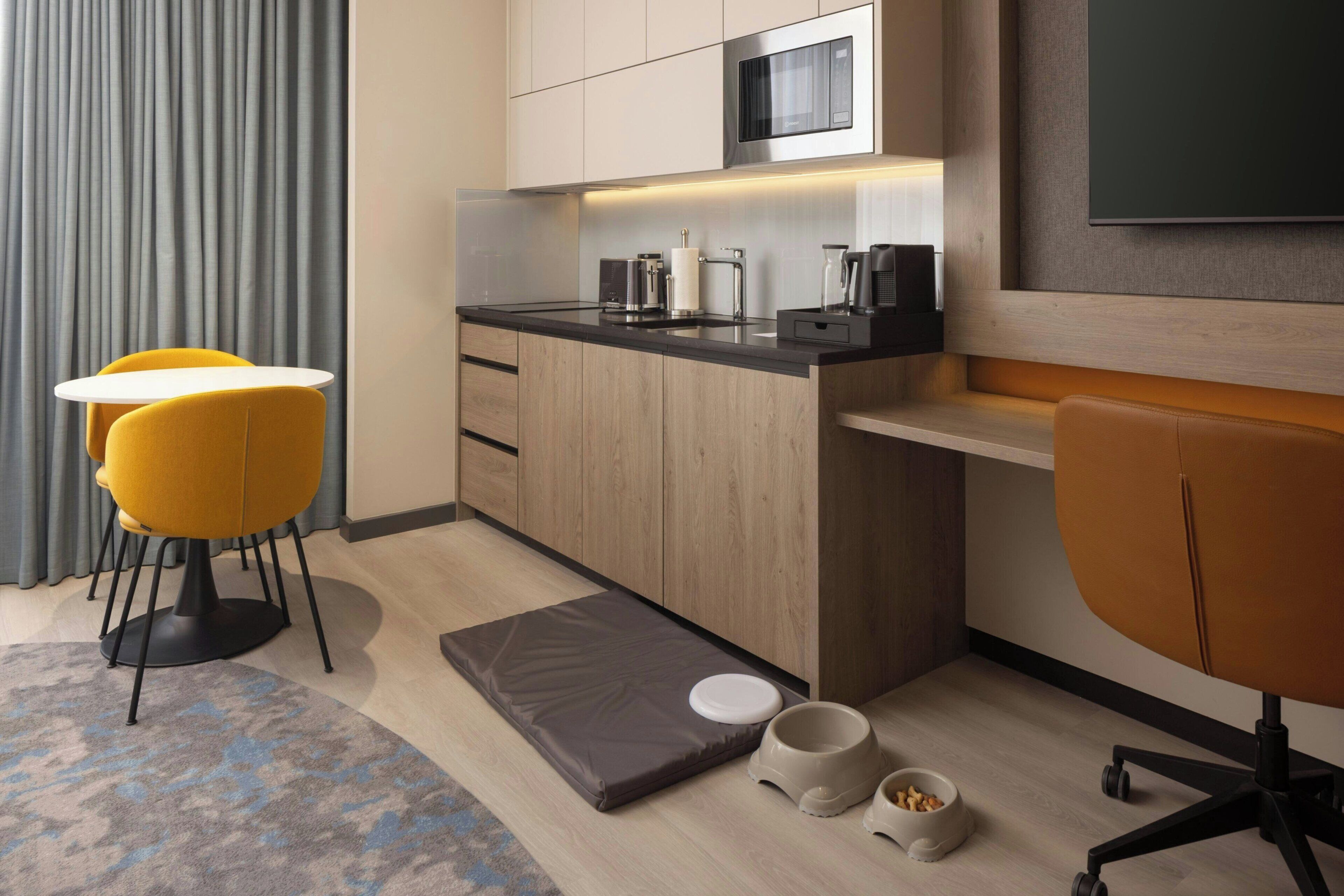 A modern kitchen with a microwave, coffee maker, and kettle, a small dining area with yellow chairs, and a pet feeding station.