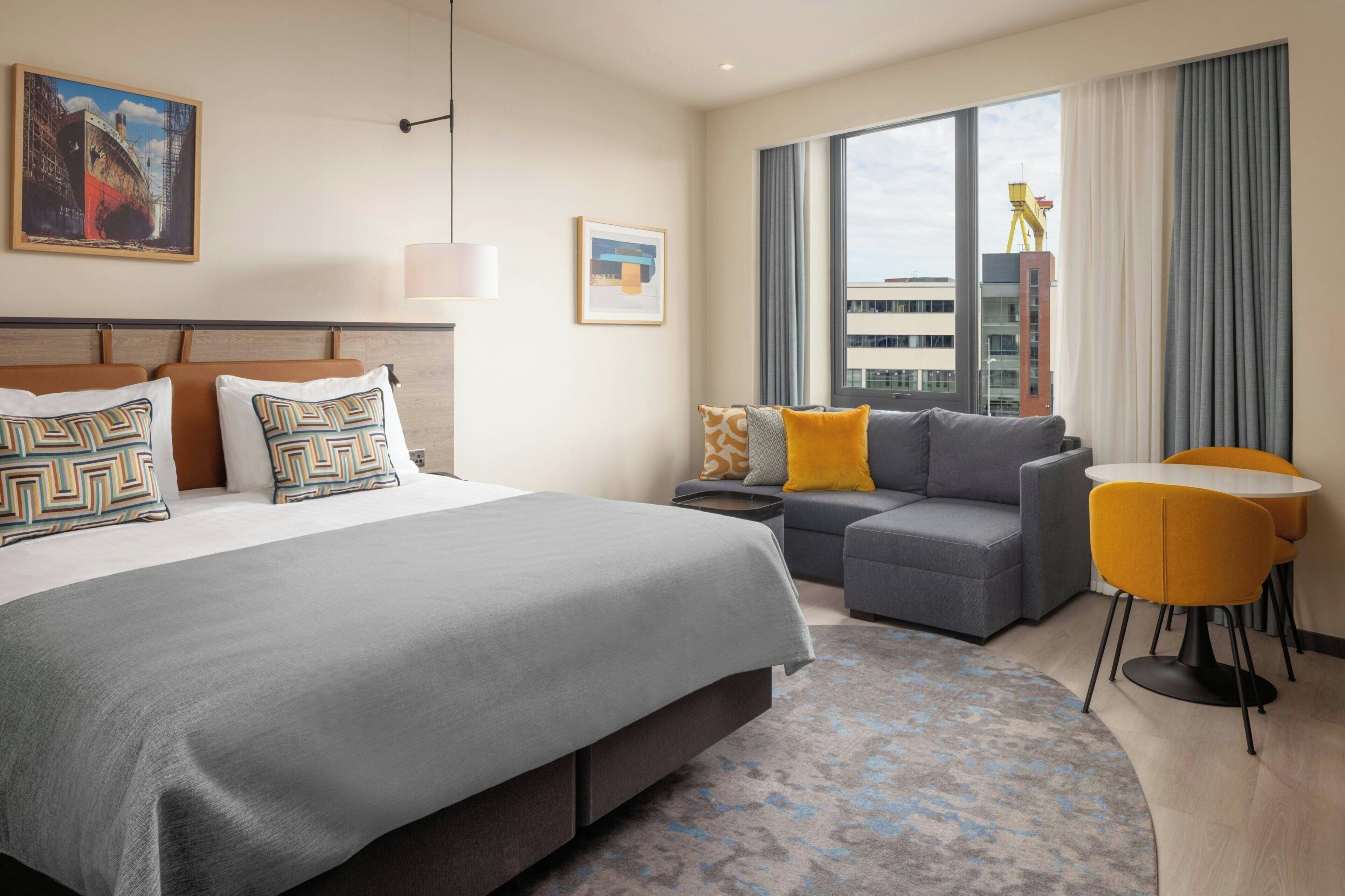 A hotel room with a large bed, a sofa, a small table, and a view of the city.