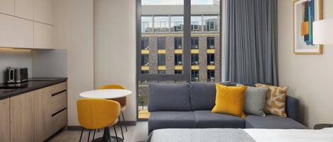 Una habitación de hotel moderna con una cama, un sofá, una mesa de comedor con sillas, una zona de cocina y un ventanal con vistas a edificios.