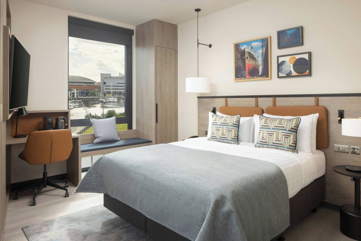 A modern hotel room with a large bed, a desk with a chair, a TV, and a view of the city.