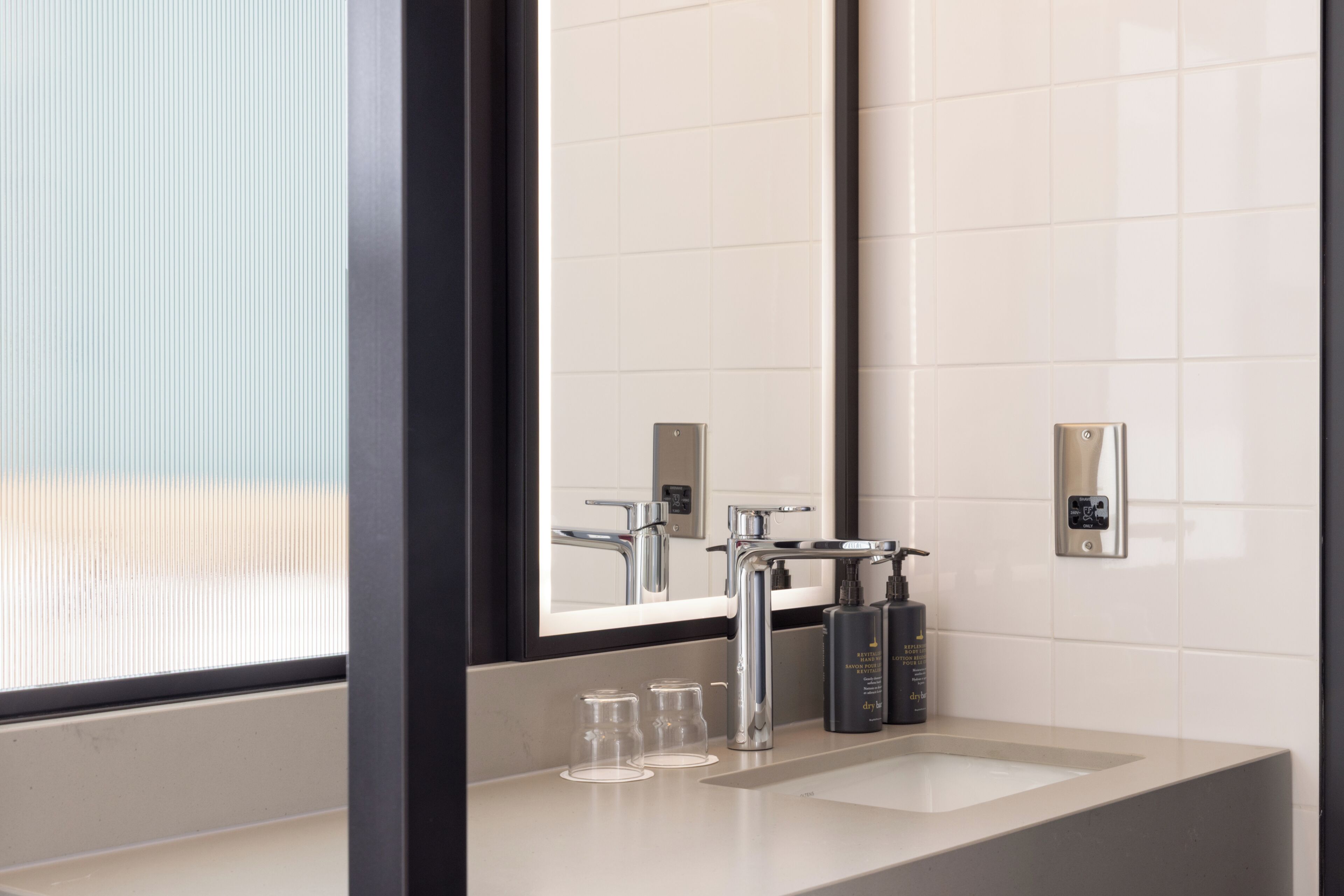 Ein modernes Badezimmer mit einem großen Spiegel, einem chromfarbenen Wasserhahn und zwei Glasflaschen auf dem Waschbecken.