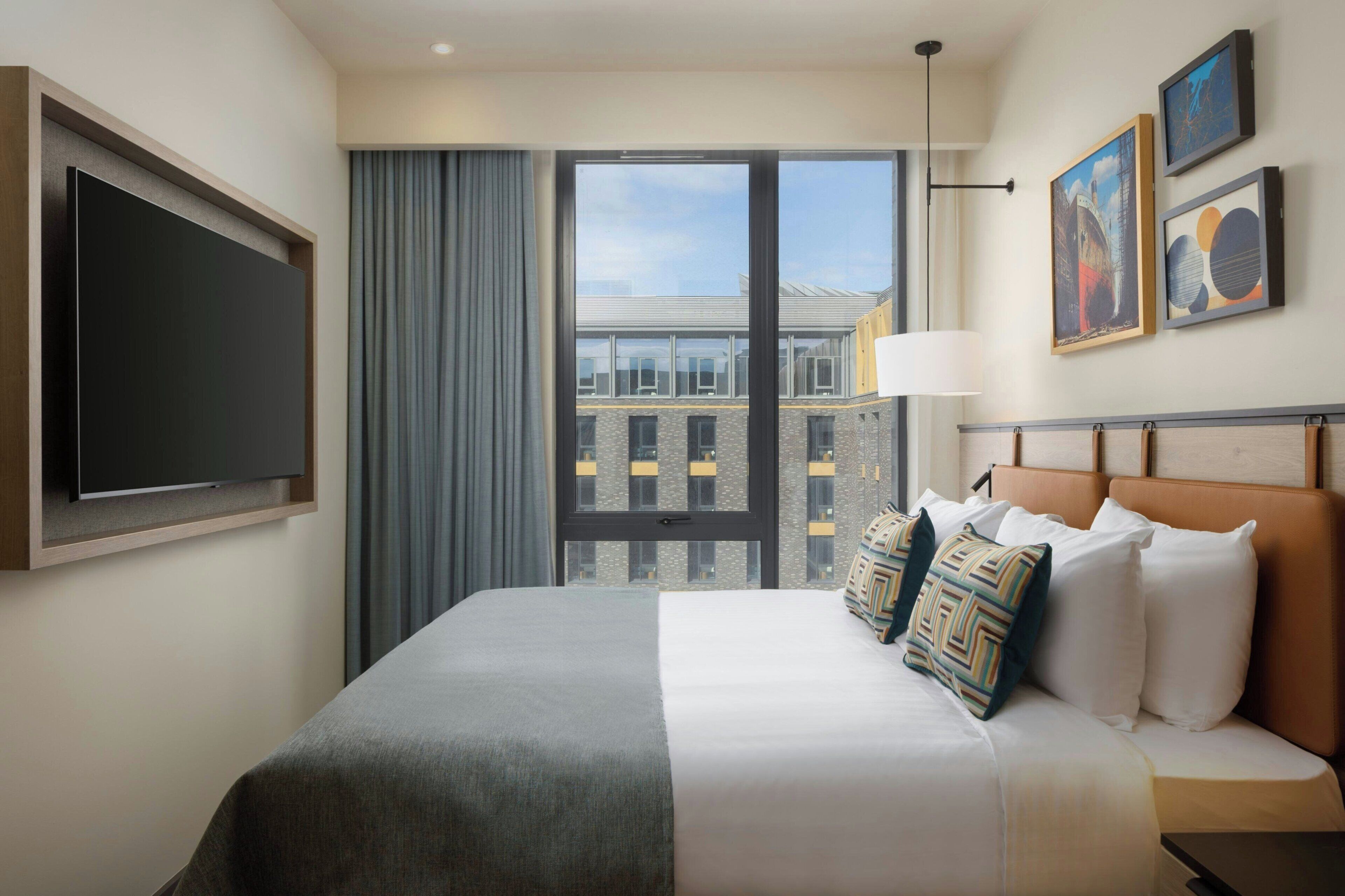 Une chambre d’hôtel avec un grand lit, un téléviseur à écran plat, une fenêtre avec des rideaux et des œuvres d’art accrochées au mur.