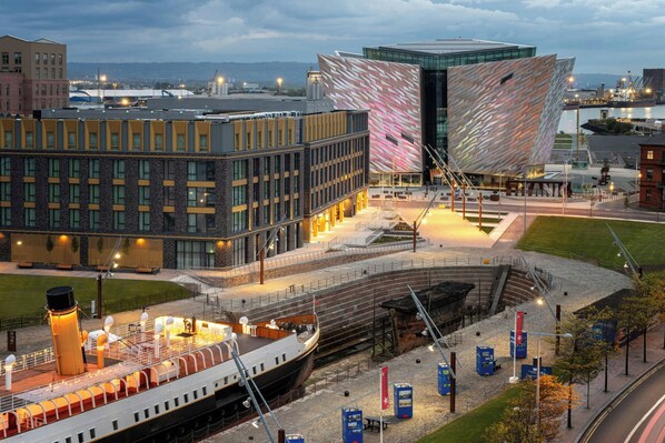 A waterfront building with a distinctive angular facade, a large open plaza, and a historic ship docked nearby. - Residence Inn By Marriott Belfast Titanic Quarter (Belfast)