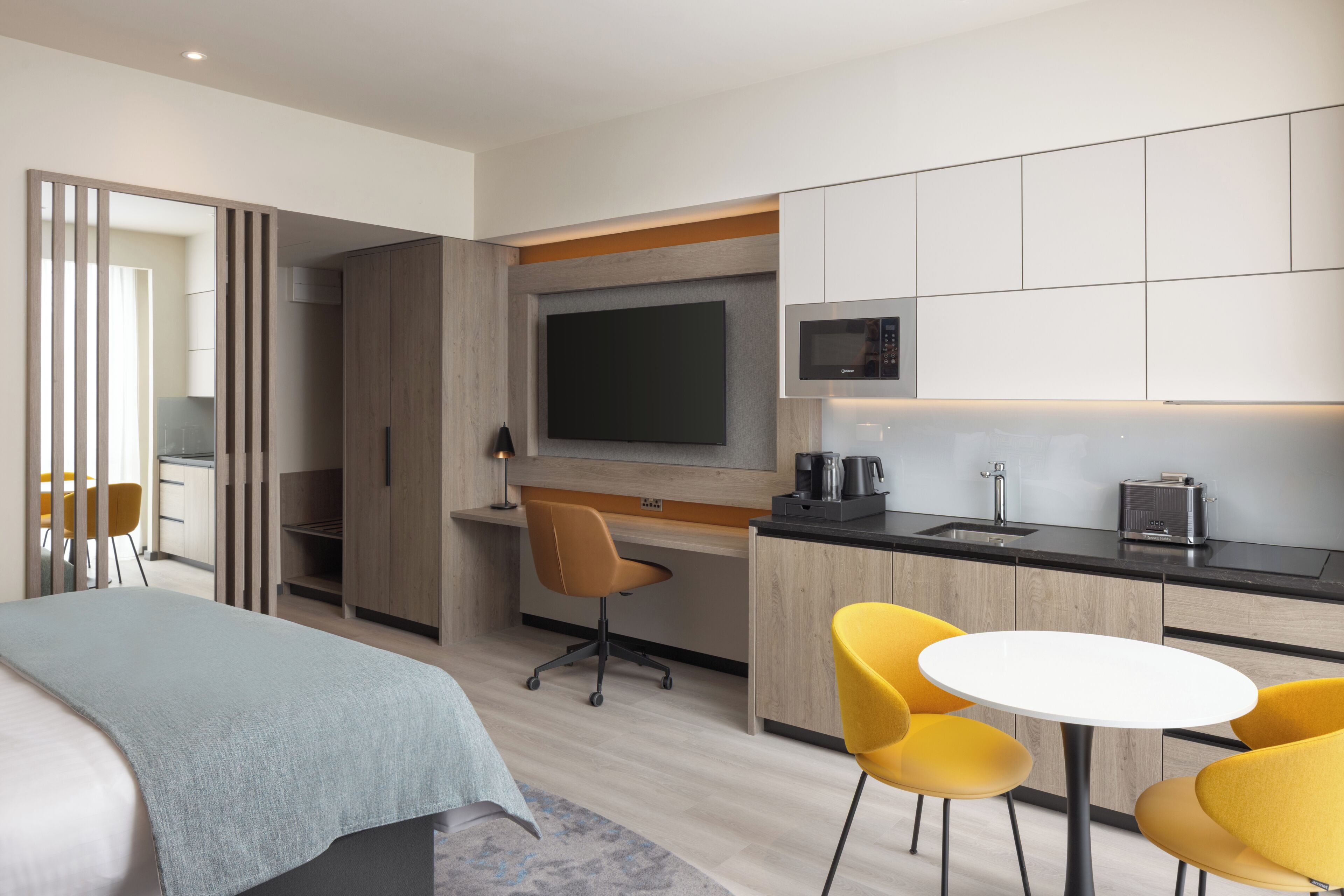 Habitación de hotel moderna con cama, escritorio, televisor, microondas y zona de comedor con sillas.