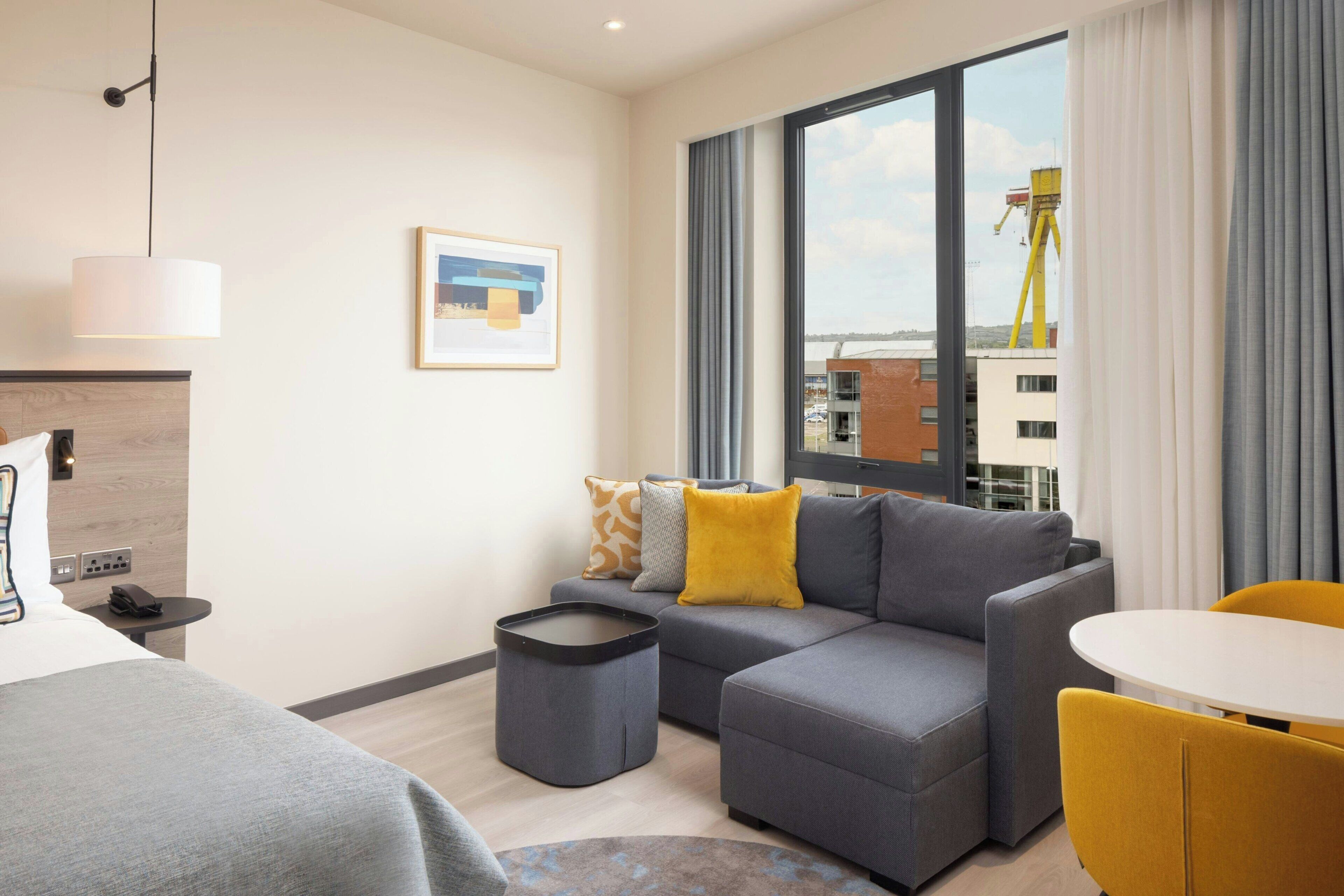 A modern hotel room with a grey sofa, a small round table, a bed, and a large window with curtains.