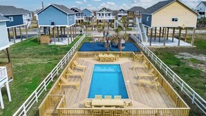 Ett poolområde med trädeck och sittplatser samt en central pool, omgivet av strandhus med upphöjda terrasser.
