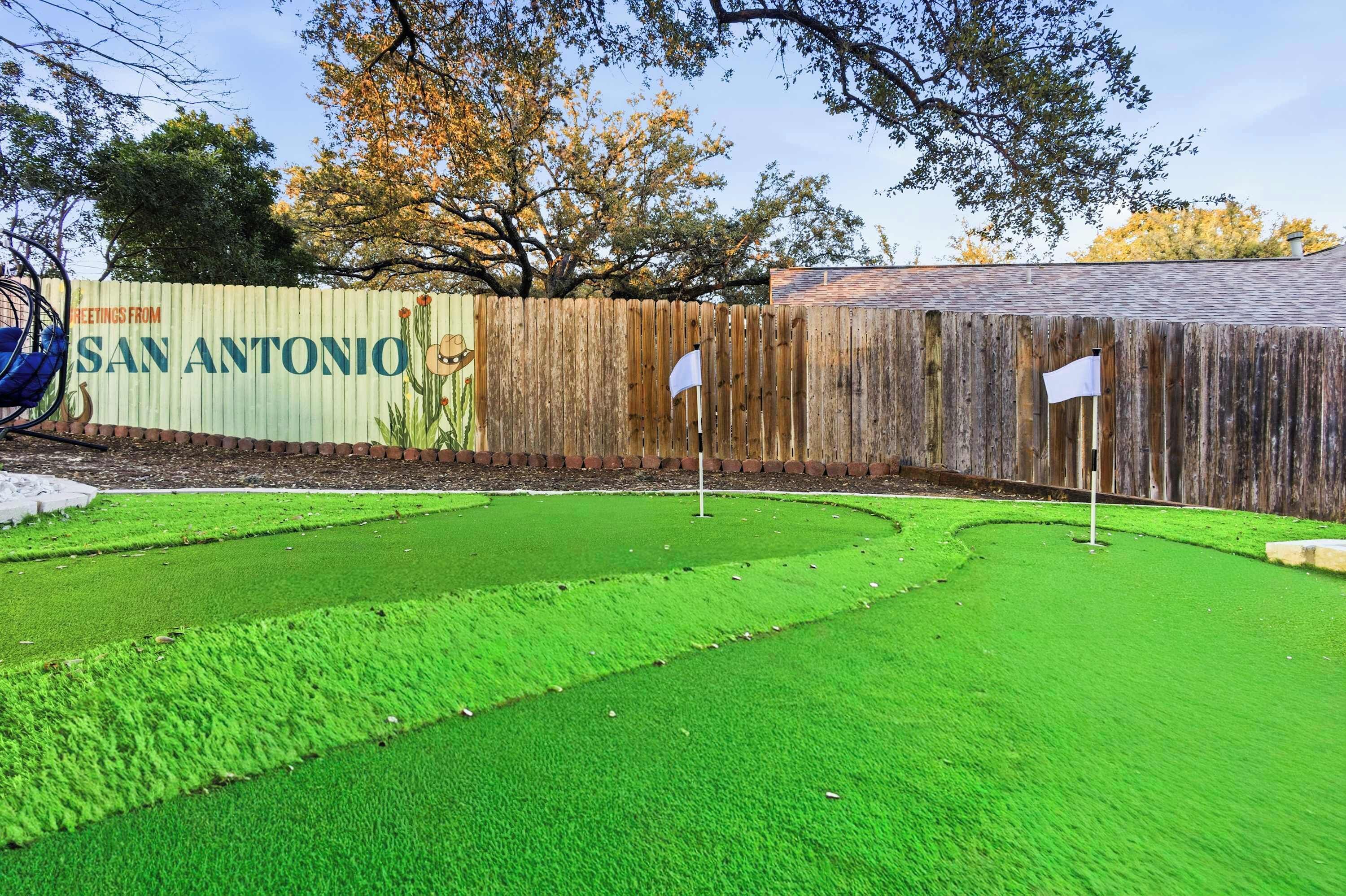 A backyard with artificial green grass, a wooden fence, and a sign that reads "Greetings from San Antonio".