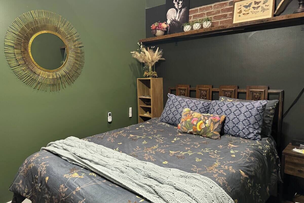 A bedroom with a bed, a round mirror, a wooden shelf with decorative items, and a brick accent wall.