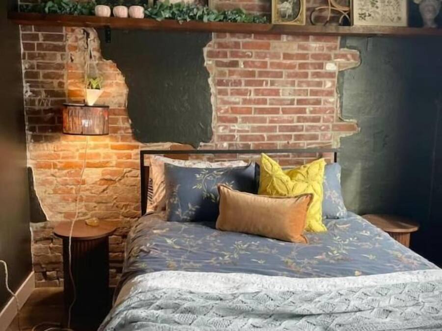 A bedroom with a brick wall, a bed with patterned bedding, and a shelf with decorative items.