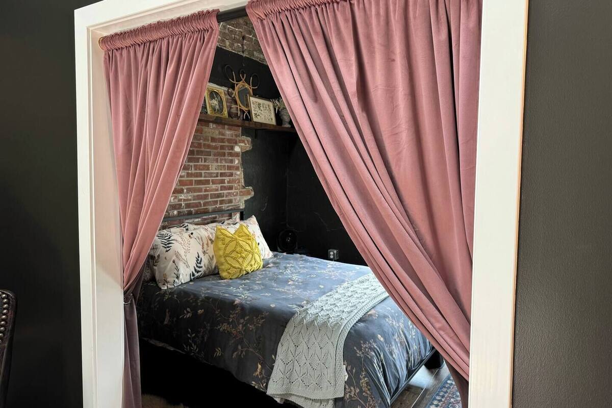 A bedroom with a bed, pink curtains, a brick wall, and a shelf with decorative items.
