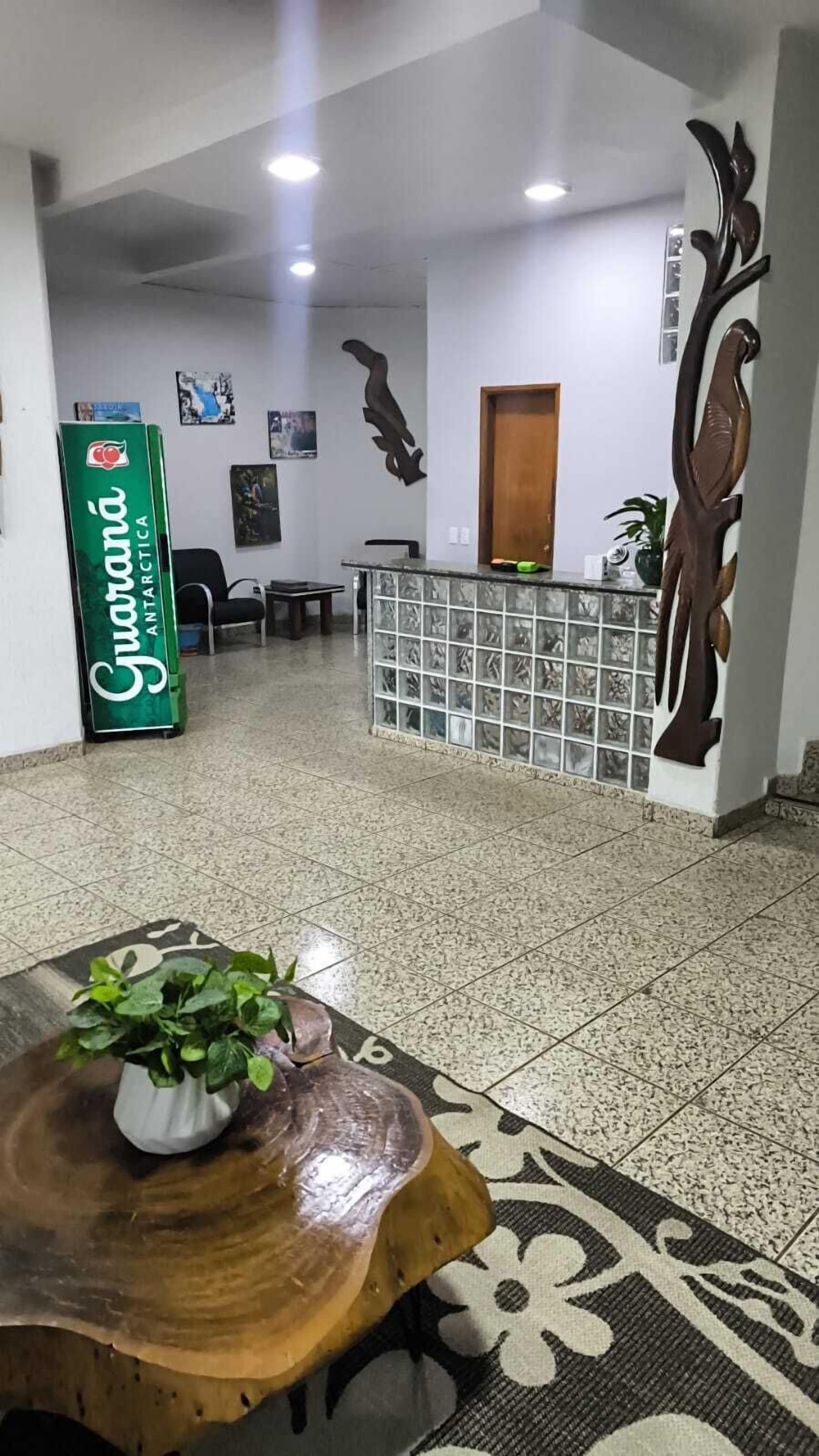 Un área de recepción con mostrador, un letrero verde, una mesa de madera con una planta y una alfombra estampada.