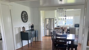 A spacious living room with a dark wooden coffee table, a gray sofa with a plaid cushion, and a dining area with a wooden table and chairs. A kitchen with a stainless steel refrigerator and a microwave is visible in the background.