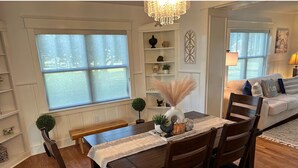 A dining area with a wooden table and chairs, a chandelier, and a view of the outdoors through large windows.