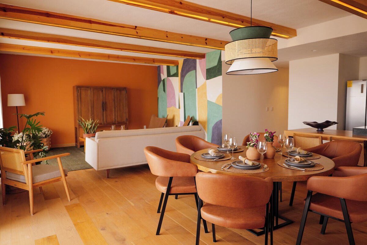 A dining area with a round table set for six, surrounded by six chairs. There is a sofa and a cabinet in the background.