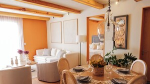 Uma sala de jantar com uma mesa de madeira posta para seis pessoas, rodeada por cadeiras de vime. Há um sofá branco e um abajur ao fundo.