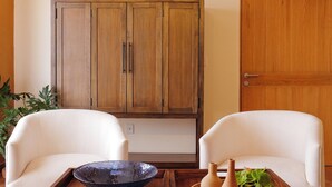 Quarto com duas cadeiras brancas, um armário de madeira e uma mesa de madeira com uma tigela e vasos.