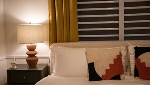 Una cama bien hecha con ropa blanca y dos almohadones decorativos, una mesita de noche con lámpara y una ventana con persianas horizontales.