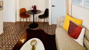 Un espacio habitable compacto con piso estampado, una mesa redonda con dos sillas, un sofá con cojines decorativos y una puerta al fondo.