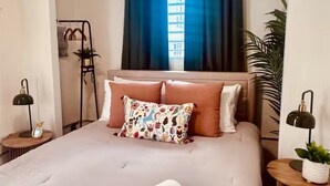 Une chambre à coucher bien rangée, avec un lit, des oreillers, une table de chevet, une lampe et une fenêtre avec des rideaux.