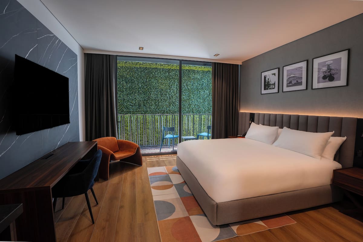 Habitación de hotel moderna con una cama grande, un escritorio, una silla, un televisor y un balcón con vistas a zonas verdes.