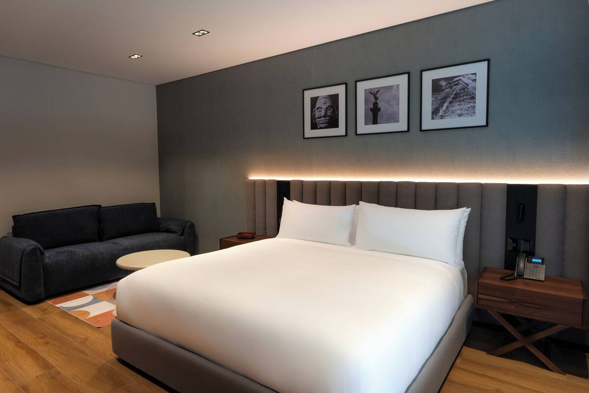 Habitación de hotel moderna con una cama grande, un sofá, una mesita de noche con un teléfono y cuadros enmarcados en la pared.