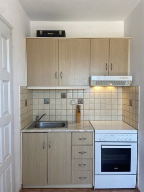 Een kleine keuken met lichte houten kastjes, een roestvrijstalen spoelbak, een witte kookplaat en een betegelde spatwand.