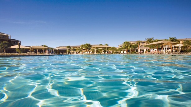 Een helder zwembad met ligbedden en parasols, omringd door resortgebouwen.