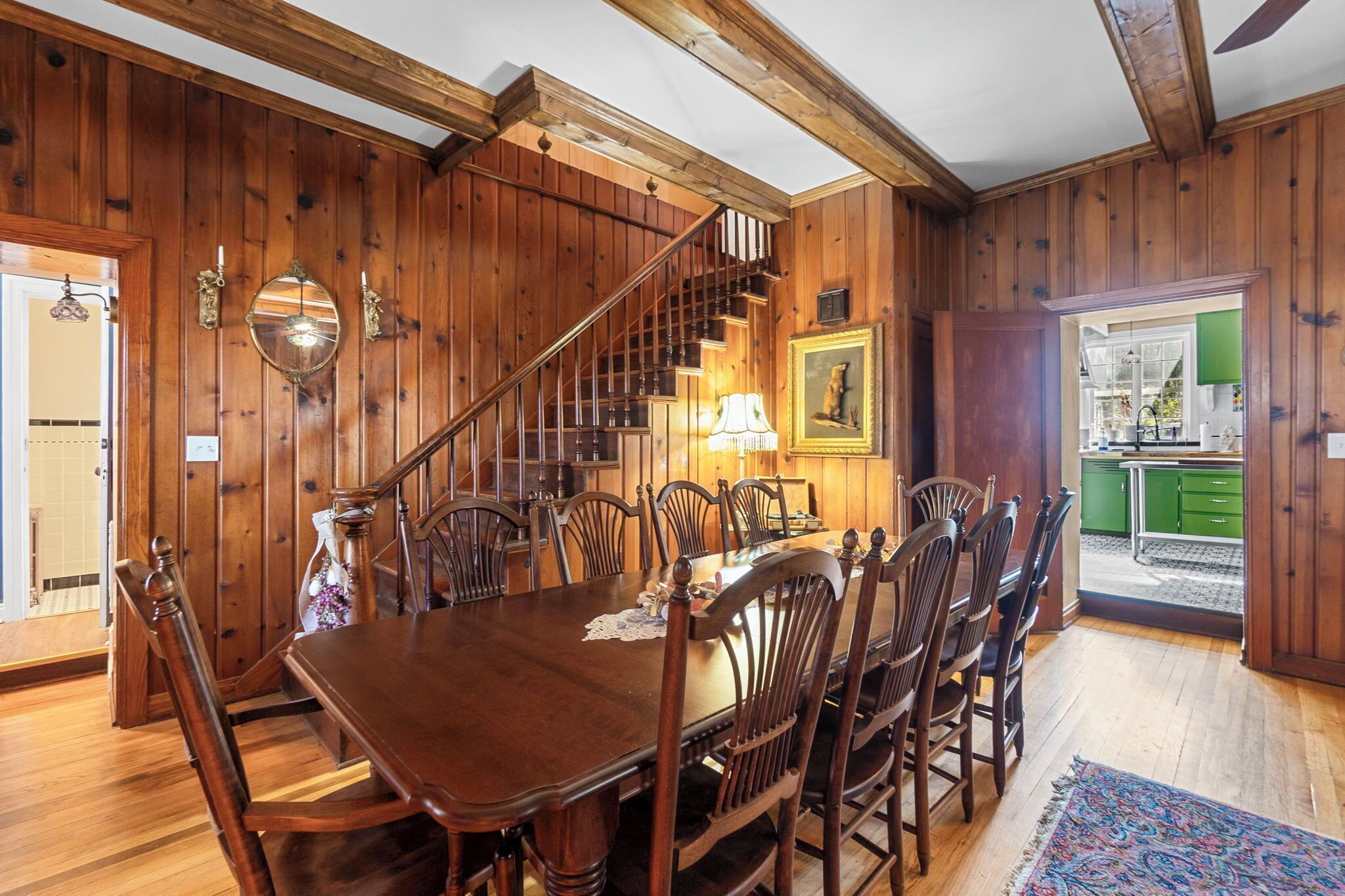 Un comedor con una mesa y sillas de madera, una escalera y una cocina visible a través de una puerta abierta.