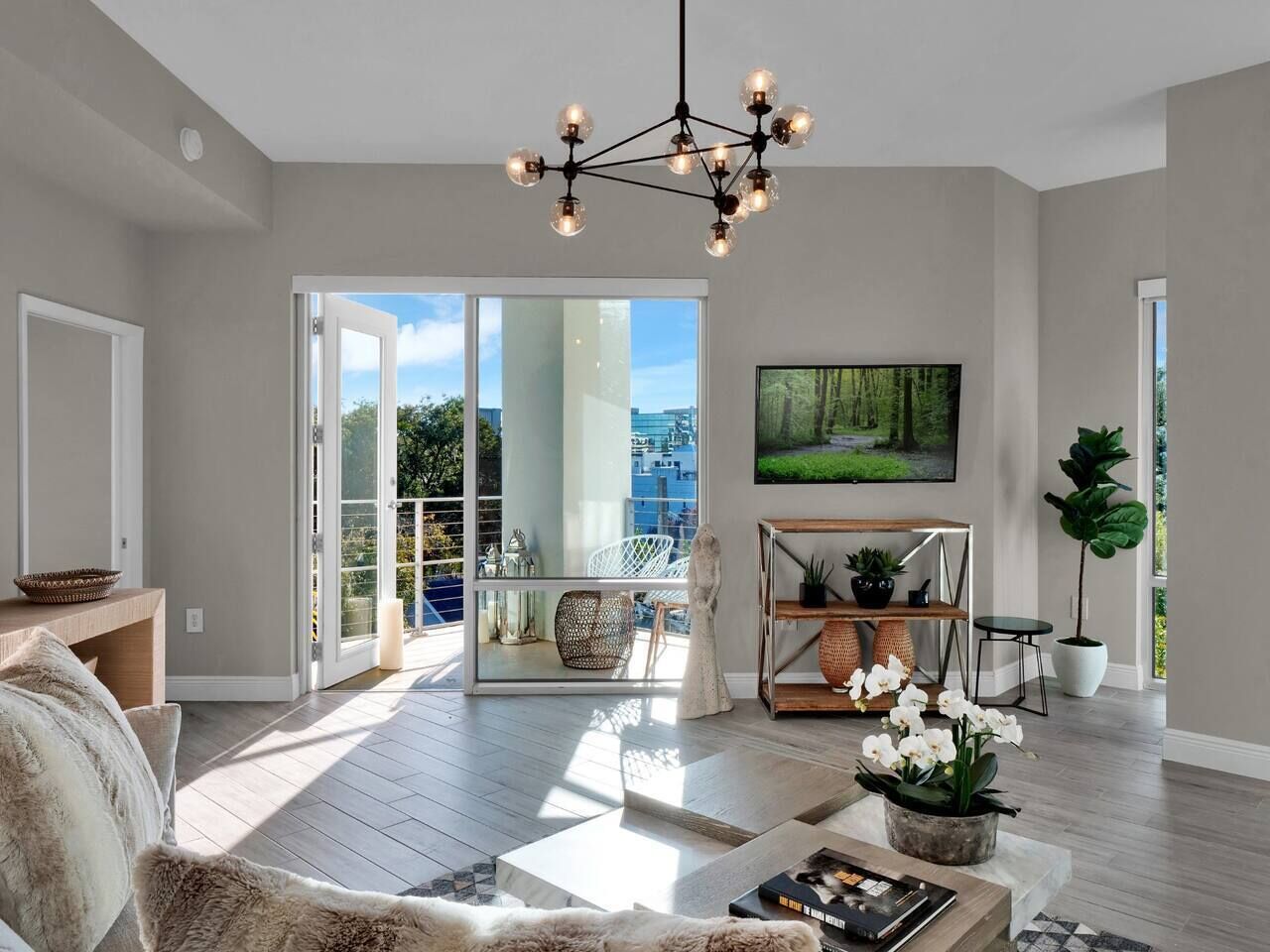 Ein geräumiges Wohnzimmer mit einem modernen Kronleuchter, einem großen Flachbildfernseher und einem Balkon mit Blick auf die Stadt.