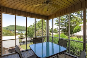 Un patio cubierto con una mesa y sillas, amplias ventanas que ofrecen vistas a un lago y colinas.