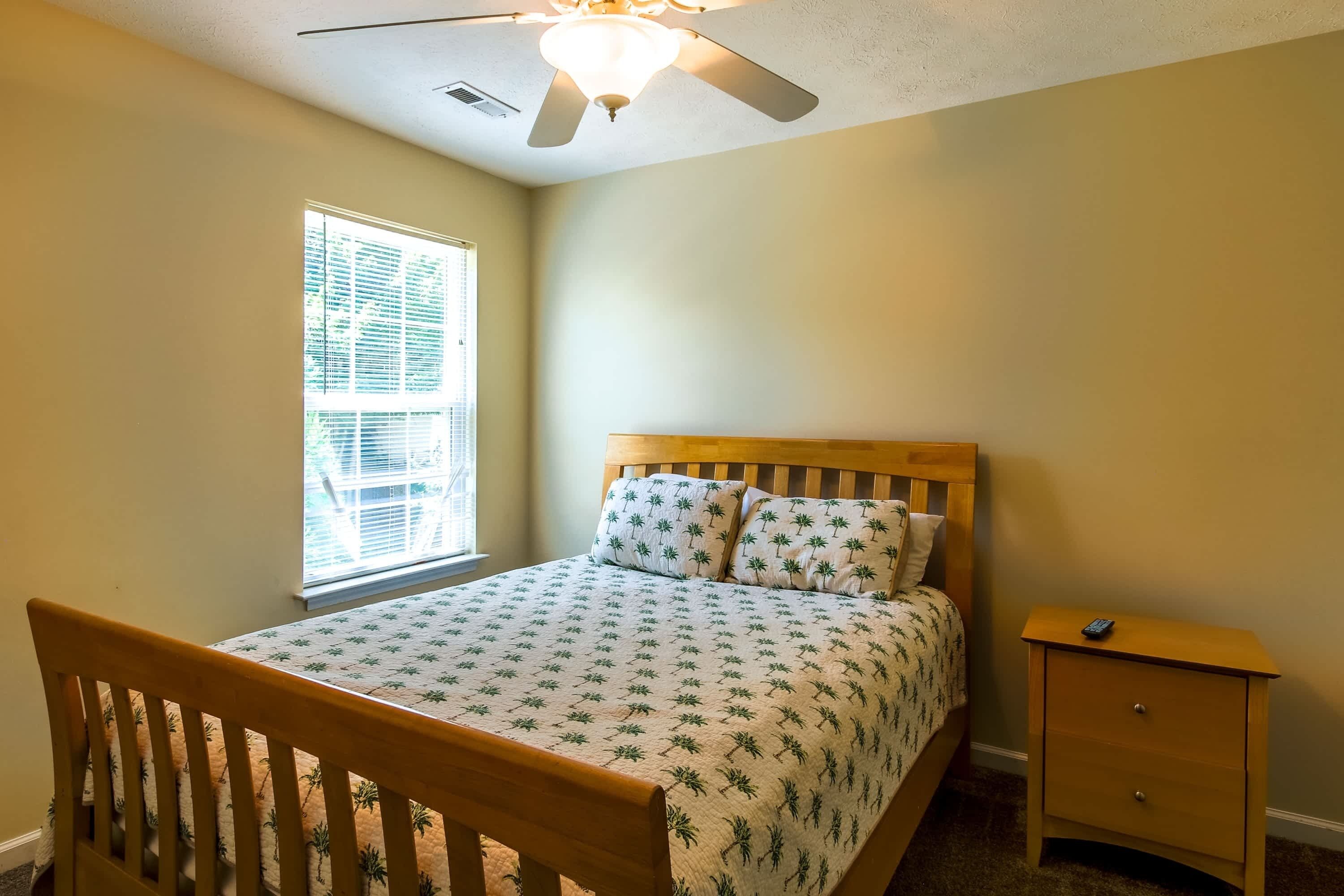 A bedroom with a bed, a nightstand, a window with blinds, and a ceiling fan.