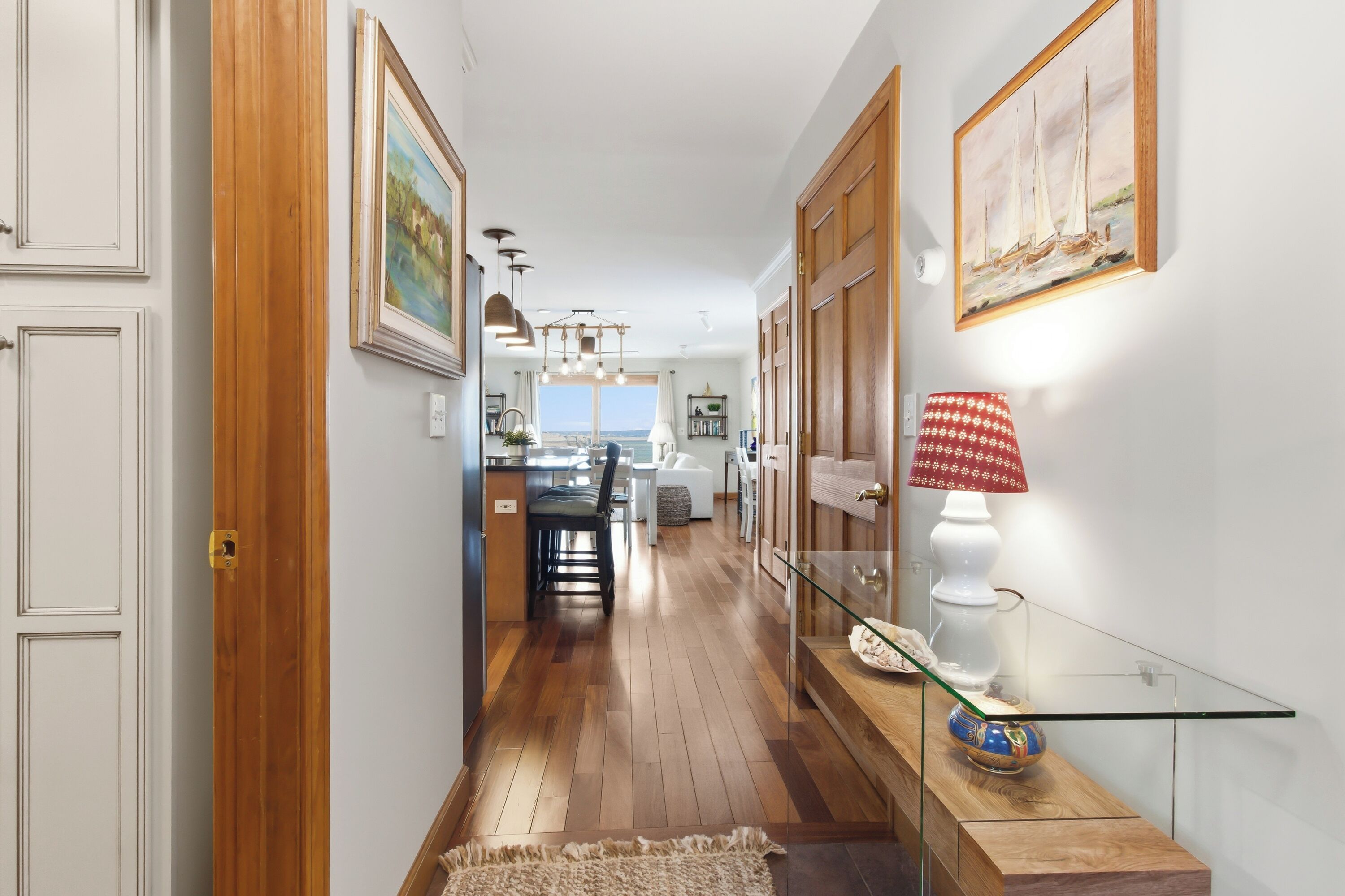 A hallway with wooden flooring, a glass table with a decorative bowl, a red patterned lamp, and framed artwork on the walls.