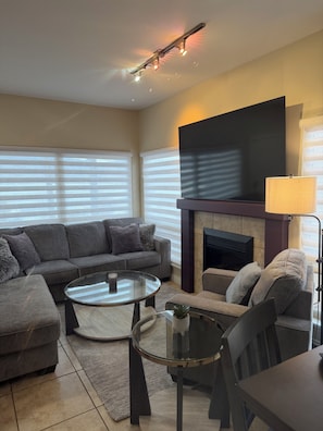A modern living room with a sectional sofa, a glass coffee table, a fireplace, and a flat-screen TV.