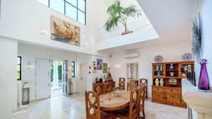 A spacious dining area with a high ceiling, a wooden dining table surrounded by chairs, and a view of the outdoors through large windows. - Casa Charador - Luxury Golf Villa (Alvor)