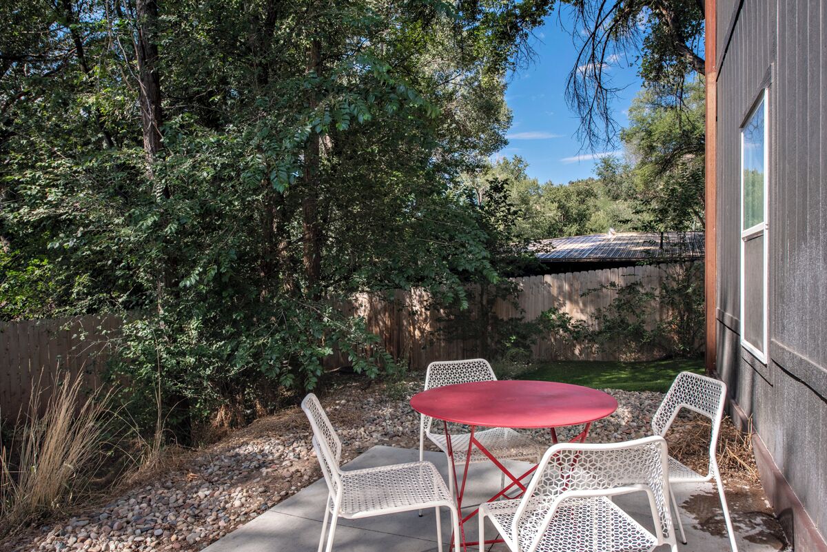 An outdoor patio with a red table and white chairs, surrounded by trees and a stone wall.