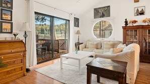 A spacious living room with a large sliding glass door leading to an outdoor area, a white coffee table, a sofa with 'ho ho ho' pillows, and a wooden cabinet. - Cozy 3BR Knotty Pine, Near Lake & Trails w Deck (Big Bear Lake)