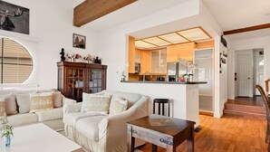 A cozy living room with a sofa, a coffee table, and a wooden table. There is a kitchen area with a refrigerator and a window. - Cozy 3BR Knotty Pine, Near Lake & Trails w Deck (Big Bear Lake)