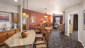 Camera d'albergo con angolo cottura, tavolo da pranzo e sedie. Nell'area cucina si nota un ampio armadio in legno, una macchina da caffè e un vaso. La zona pranzo presenta un tappeto decorato e una ventola a soffitto.