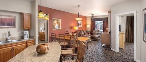 A hotel room with a kitchen area, dining table, and chairs. There is a large wooden cabinet, a coffee maker, and a vase on the kitchen counter. The dining area has a patterned rug and a ceiling fan.
