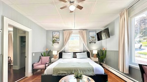 Une chambre à coucher comprenant un lit, des tables de chevet, un ventilateur de plafond, une télévision et une fenêtre avec des rideaux.