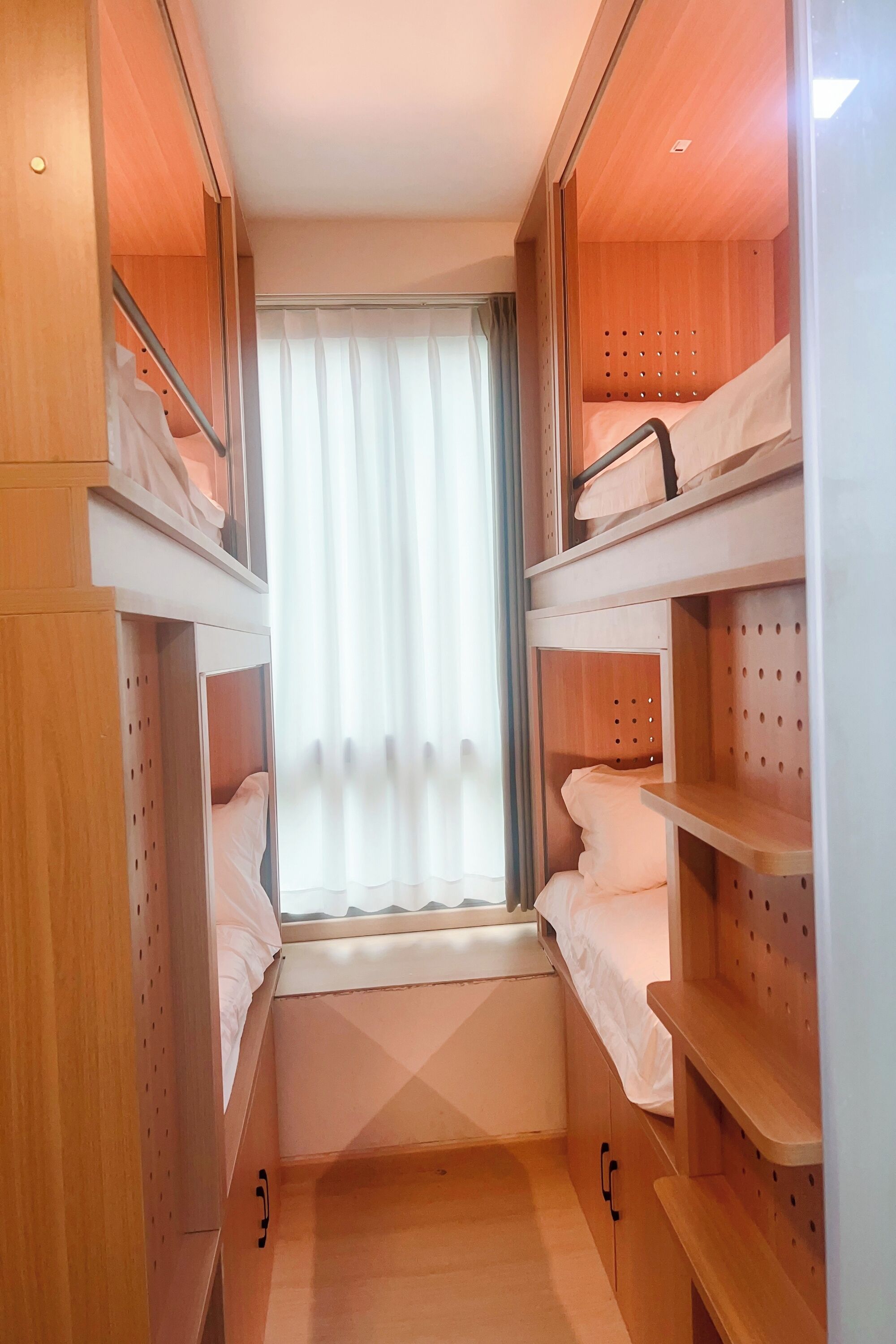 Een compacte kamer met ingebouwde houten planken, een raam met gordijnen, en een stapelbed.
