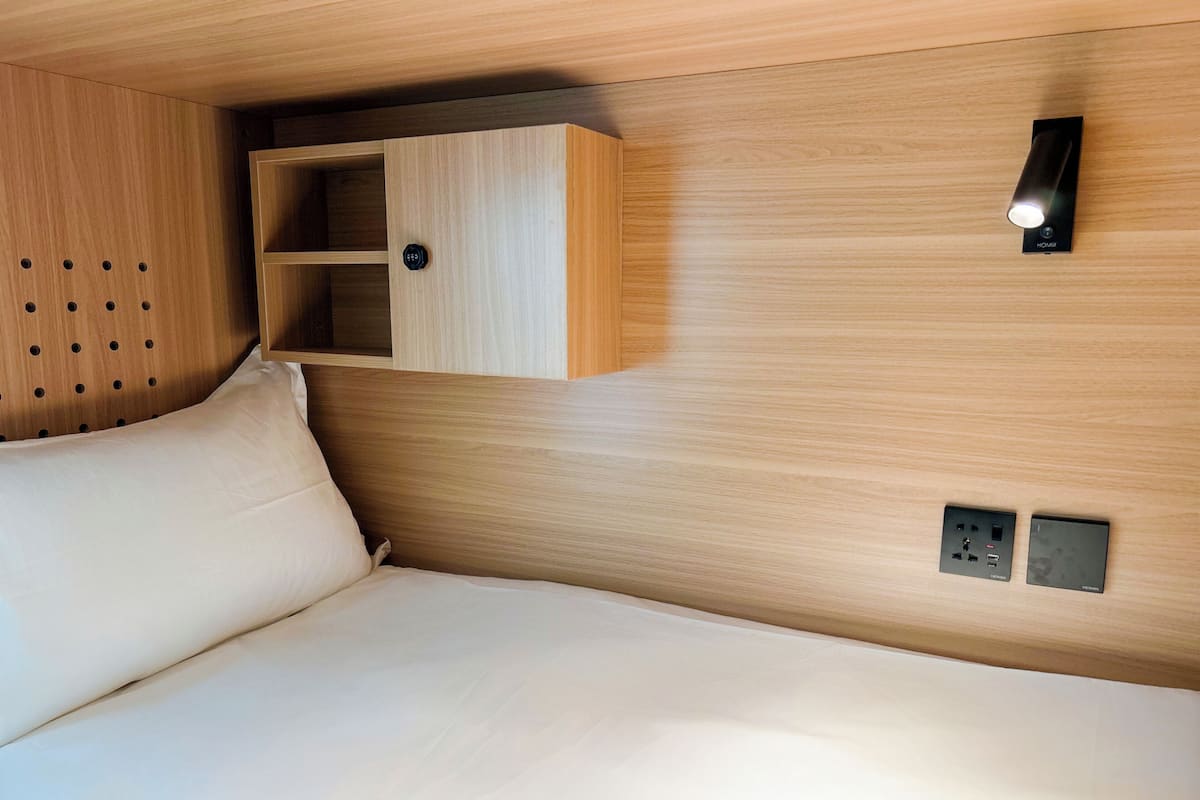 A bed with a headboard, a wooden shelf unit, a wall-mounted light, and electrical outlets.