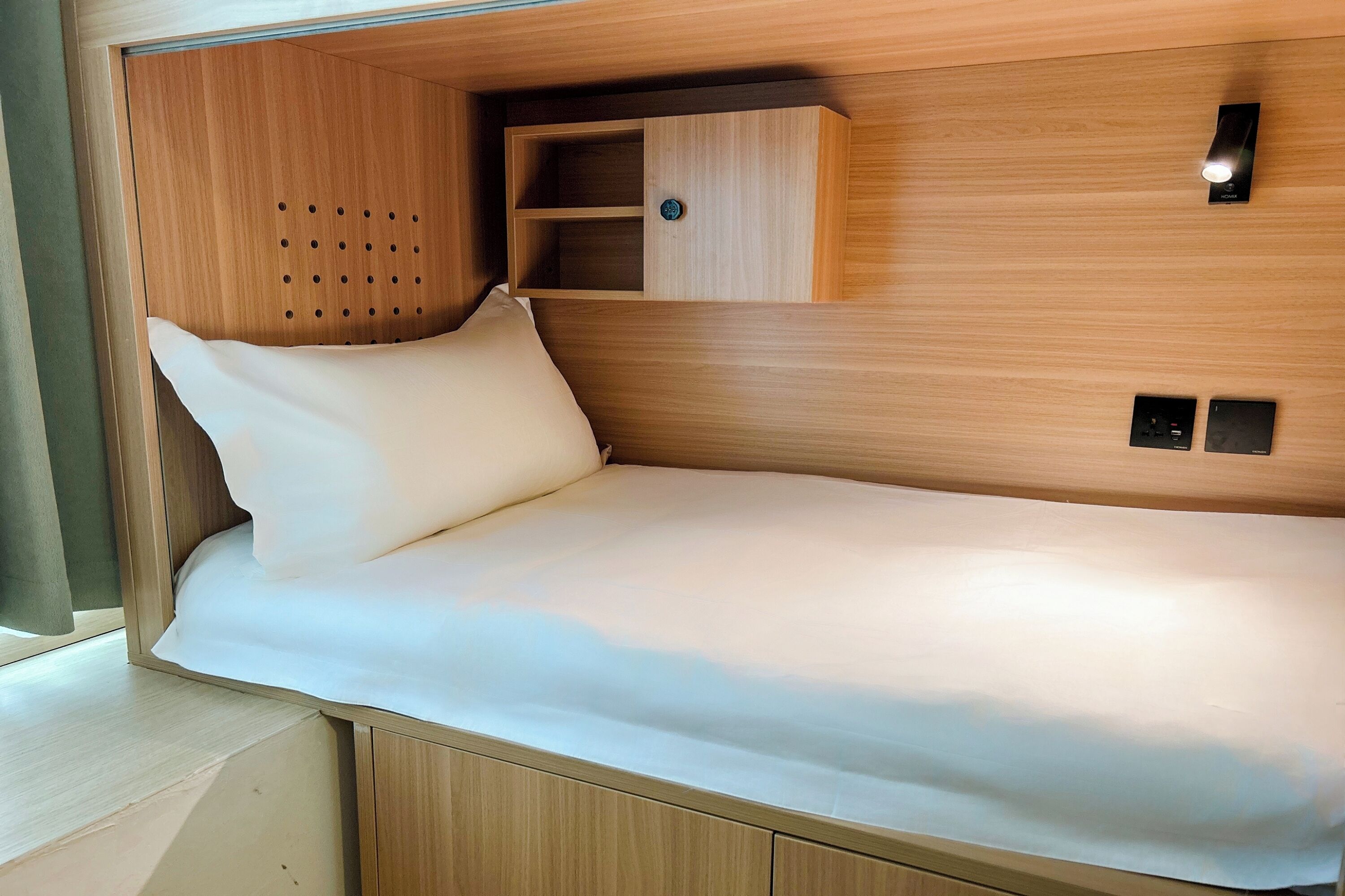 Un lit compact doté d’une tête de lit en bois, d’une petite étagère et d’un luminaire fixé au mur.