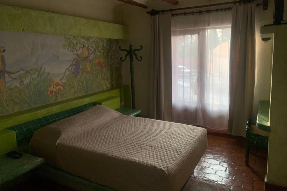 Une pièce comprenant un lit, un fauteuil vert, un mur orné d’une fresque et une fenêtre avec des rideaux.