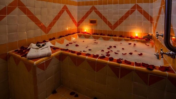 A bathtub with rose petals and candles, surrounded by patterned tiles.