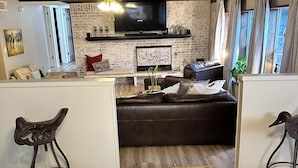 A living room with a brown leather sofa, a TV mounted on a brick wall, and a small table with a plant. - Tournament Ready Stay •Close to Parks •3 bedroom/2 bath •Pool/HotTub (College Station)