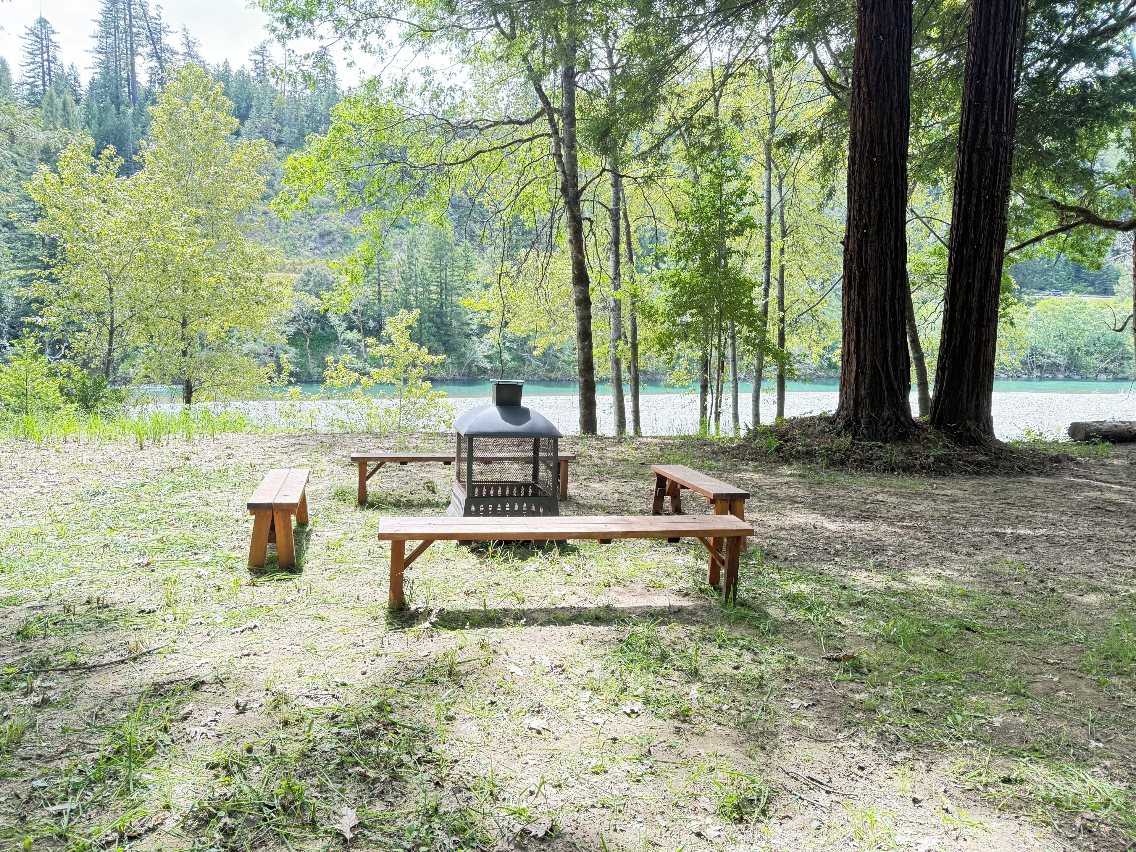 A picnic area with benches and a grill, surrounded by trees and a lake.
